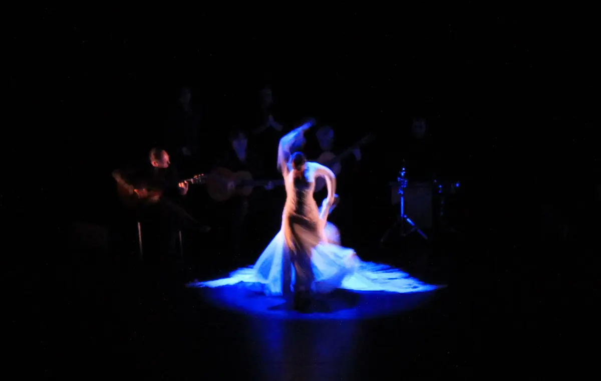 En flamencodanser i en flagrende blå kjole drejer rundt på en mørk scene oplyst i blåt, med musikere siddende bag hende