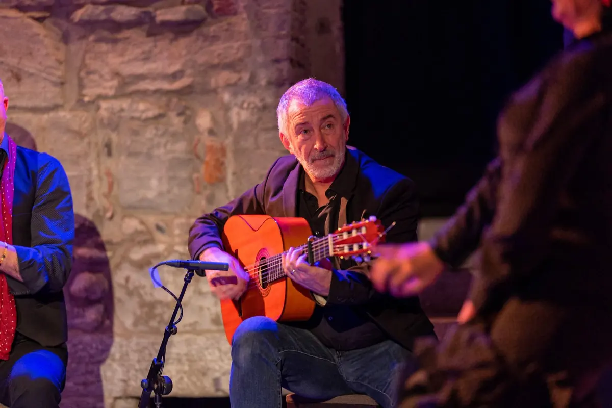En flamencoguitarist i en mørk jakke spiller på en gyldenorange guitar på en lille scene med stenvægge, med en mikrofon foran sig og en anden optrædende ved sin skulder