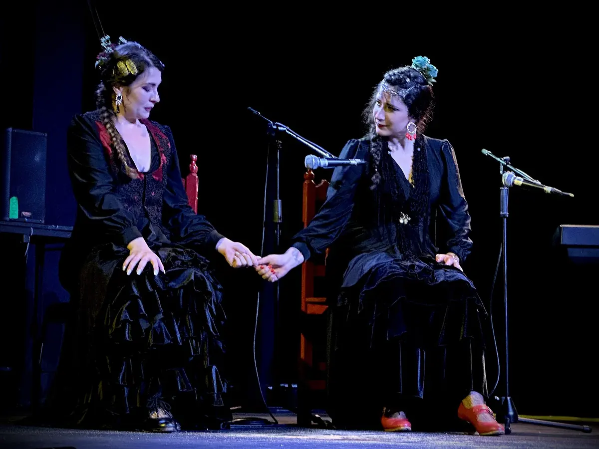 To kvinder i sorte flamencokjoler sidder side om side på en mørk scene og holder hinanden i hånden, hver med en mikrofonstander foran sig