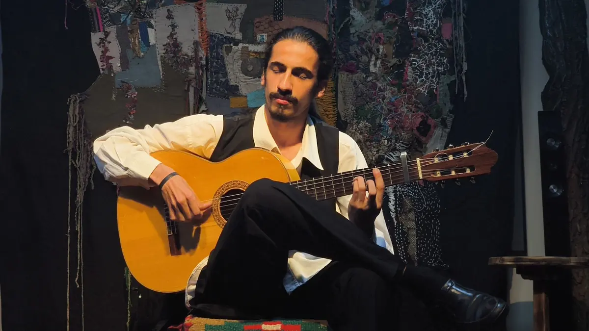 En flamencoguitarist i hvid skjorte og sort vest sidder med det ene ben over det andet og guitaren holdt næsten vandret, spillende foran et mønstret stofbagtæppe